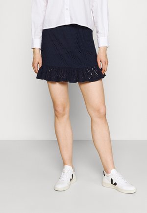 Person wearing a white long-sleeve shirt, navy blue eyelet mini skirt with ruffle hem, and white sneakers with black accents, standing on a gray floor.