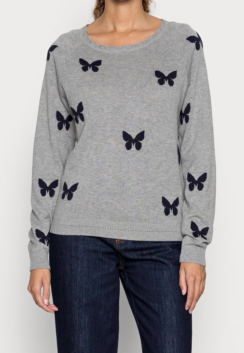 Femme portant un pull gris avec des motifs papillons noirs, accompagné d'un jean bleu foncé, debout devant un fond clair uni.