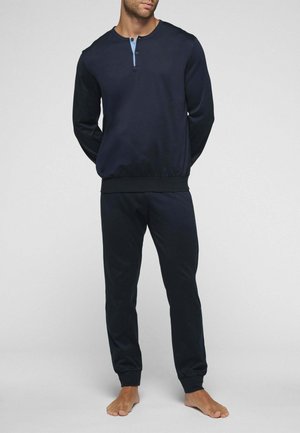 Uomo che indossa una maglia henley a maniche lunghe blu navy e pantaloni da jogging abbinati, in piedi a piedi nudi su sfondo grigio chiaro.