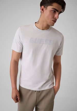 Young man with dark hair wearing a light grey GUESS T-shirt and beige pants, standing with one hand in his pocket, looking to his side.