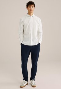 Balts garais krekls ar garām piedurknēm un gaišām vertikālām svītrām, apvienots ar tumši zilām biksēm un baltām sporta kurpēm ar pelēkiem akcentiem. Gluda auduma tekstūra.