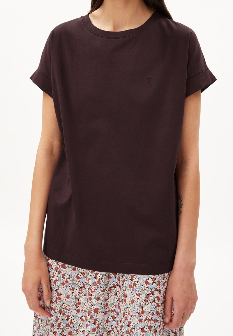 Femme portant un t-shirt manches courtes marron foncé avec des manches retroussées et une jupe à motif floral avec de petites fleurs blanches et rouges.