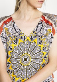 Blouse à col en V présentant un motif floral complexe jaune et noir avec des accents rouges, fabriquée en tissu léger.