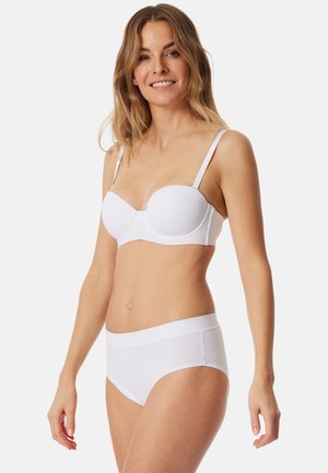 Lachende vrouw met golvend blond haar, gekleed in een witte strapless beha en witte hoge tailleonderbroek, staand tegen een effen achtergrond.