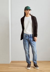 Zwarte gebreide cardigan over een wit T-shirt, blauwe spijkerbroek en donkere sneakers. De pet is groen, wat de casual outfit compleet maakt.