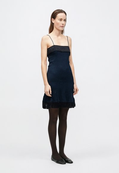 Tengerészkék testhezálló ruha vékony pántokkal, texturált anyagból, rojtos díszítéssel a nyakkivágásnál és az alján. Fekete harisnyával és lapos talpú cipővel párosítva.