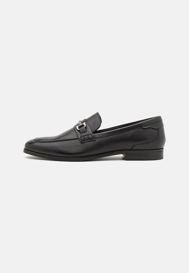 Mocassin pour homme en cuir noir avec détail en métal argenté, petit talon et bout carré arrondi, présenté sur un fond blanc.