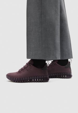 Zapatillas deportivas de color púrpura oscuro con cordones y suelas gruesas y ranuradas, llevadas con calcetines negros y pantalones grises de pierna ancha sobre un fondo liso.