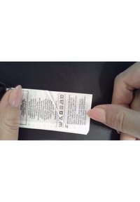 Skötselmärke med tvättinstruktioner, skötsel-symboler och flerspråkig text på en vit tygetikett som hålls mot en mörk bakgrund.