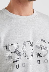 Närbild av en person som bär en ljusgrå skjorta med en pixelartad "BOSS"-design och texten "HUGO BOSS" nedanför.