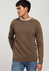 Brązowy sweter z dzianiny z okrągłym dekoltem i prążkowanymi mankietami. Charakteryzuje się teksturowaną powierzchnią i luźnym krojem. Noszony na białej koszuli.