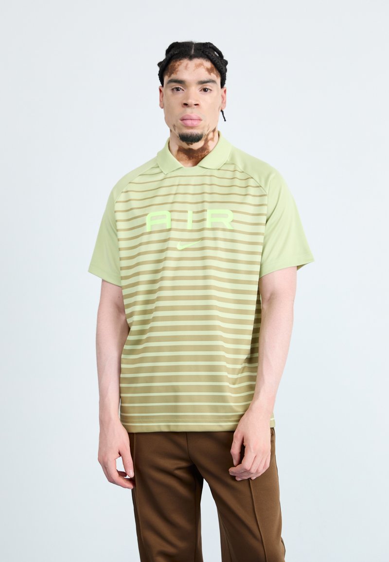 Polo vert rayé avec un col, présentant un grand graphique "AIR" en vert clair et un logo Nike, associé à un pantalon marron.