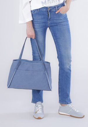 Persona con jeans blu e sneakers chiare tiene una grande borsa a tracolla blu per una cinghia al lato.