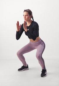 Crop top nero a maniche lunghe, leggings lavanda con strisce laterali nere e scarpe da ginnastica nere. Modella in posizione di squat, mani alzate.