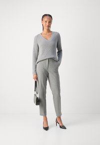 Pull gris à côtes avec un col en V, associé à un pantalon à carreaux gris clair. Le mannequin tient un petit sac à main noir et porte des talons aiguilles noirs.