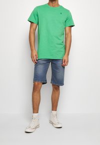 Hombre vestido con una camiseta de manga corta verde brillante, pantalones cortos de mezclilla azules hasta la rodilla, zapatillas blancas de caña alta, de pie frente a un fondo blanco.