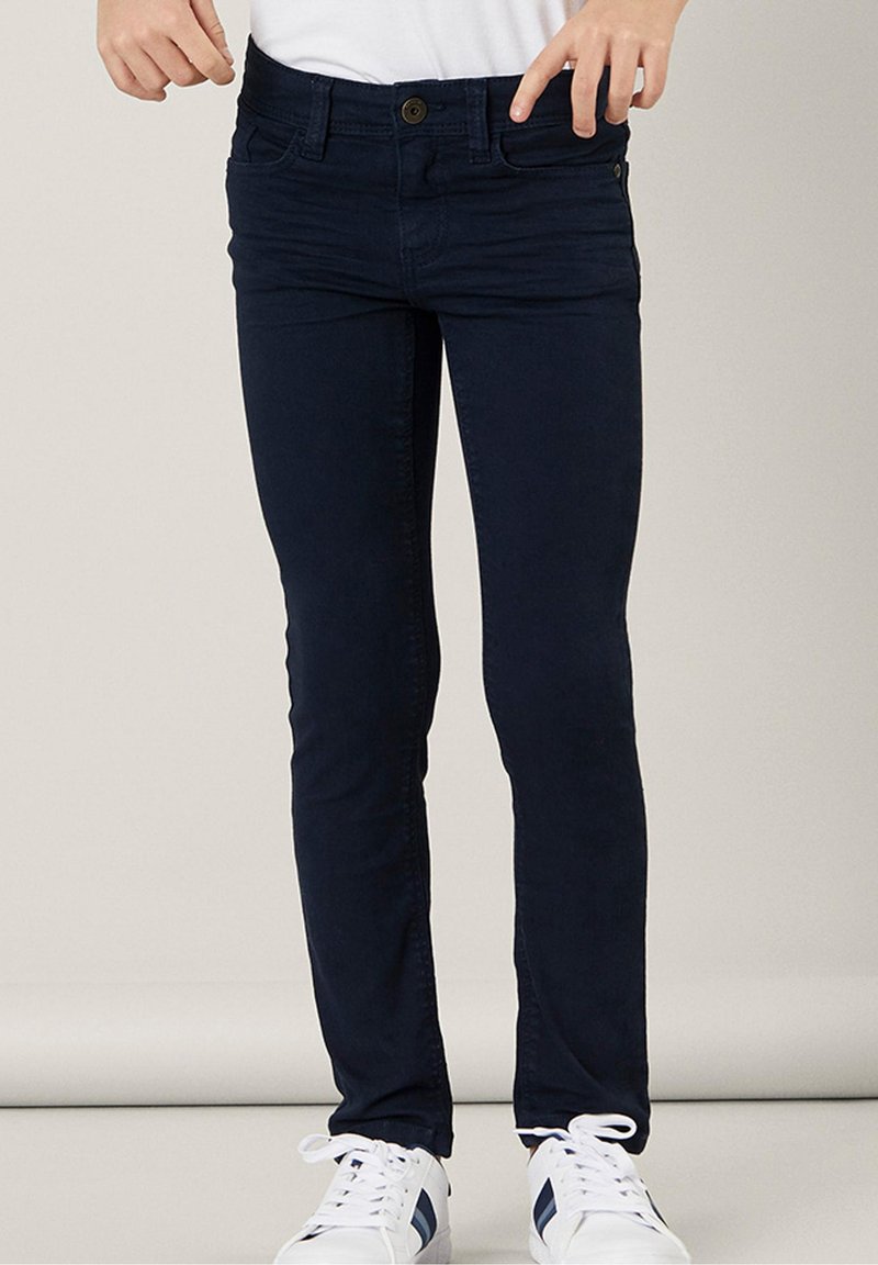 Personne portant un pantalon slim bleu foncé et des baskets blanches à rayures bleu marine, debout devant un fond clair uni.