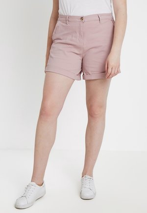 Frau trägt staubrosa Shorts mit umgeschlagenem Saum, vorderen Taschen und Knopf, kombiniert mit weißen Sneakern und einem weißen Oberteil.