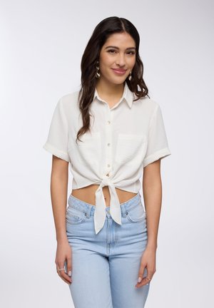 Camicia bianca a maniche corte con colletto e nodo davanti, abbinata a jeans azzurri a vita alta. Tessuto morbido con due tasche frontali.