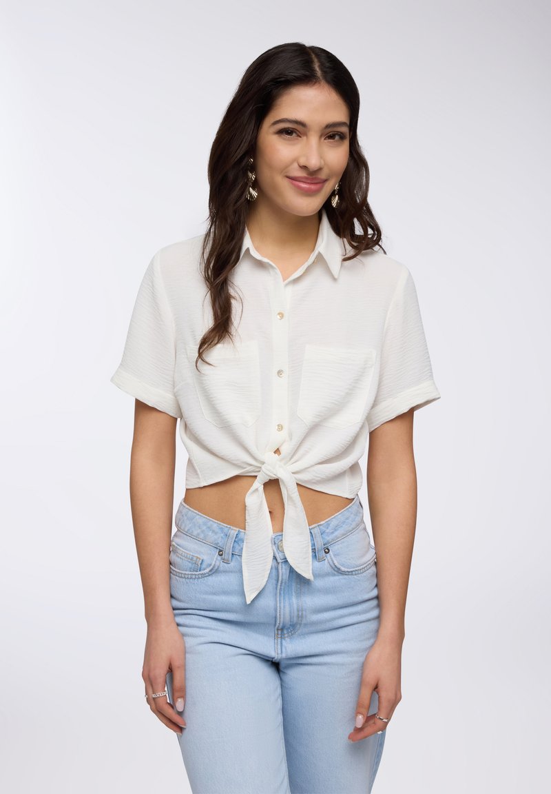Camicia bianca a maniche corte con colletto e nodo davanti, abbinata a jeans azzurri a vita alta. Tessuto morbido con due tasche frontali.