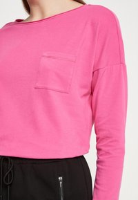 Fuchsia Langarmshirt mit rundem Halsausschnitt und einer Brusttasche; weicher Baumwollstoff; kombiniert mit schwarzen Hosen mit Reißverschlusstaschen.