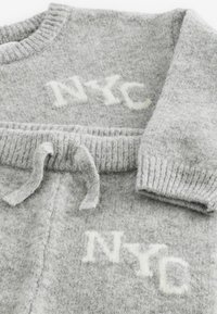 Grijze gebreide trui met geribbelde manchetten en rand. "NYC" staat prominent weergegeven in het wit, met een zachte textuur en trekkoorddetails.