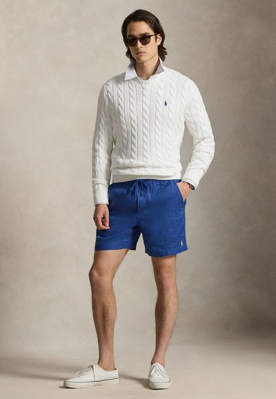 Λευκό πουλόβερ με πλέξη καλτσοδετού, μπλε σορτς με κορδόνι, ανοιχτόχρωμα παπούτσια και γυαλιά ηλίου. Η ενδυμασία χαρακτηρίζεται από casual, πολυεπίπεδη σχεδίαση.