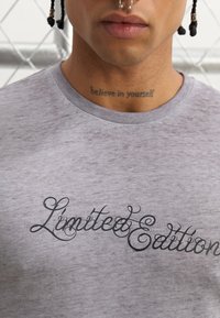 T-shirt gris à manches courtes en tissu doux, ornée du texte en cursive noire "Édition Limitée" sur la poitrine. Encolure ronde et texture subtile.
