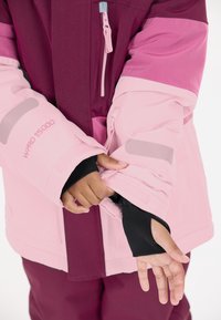 Roze en burgundy jack met contrasterende mouwen, voorzien van een rits, logo en duimgaten in de mouwen. Het materiaal is glad, waarschijnlijk waterdicht.