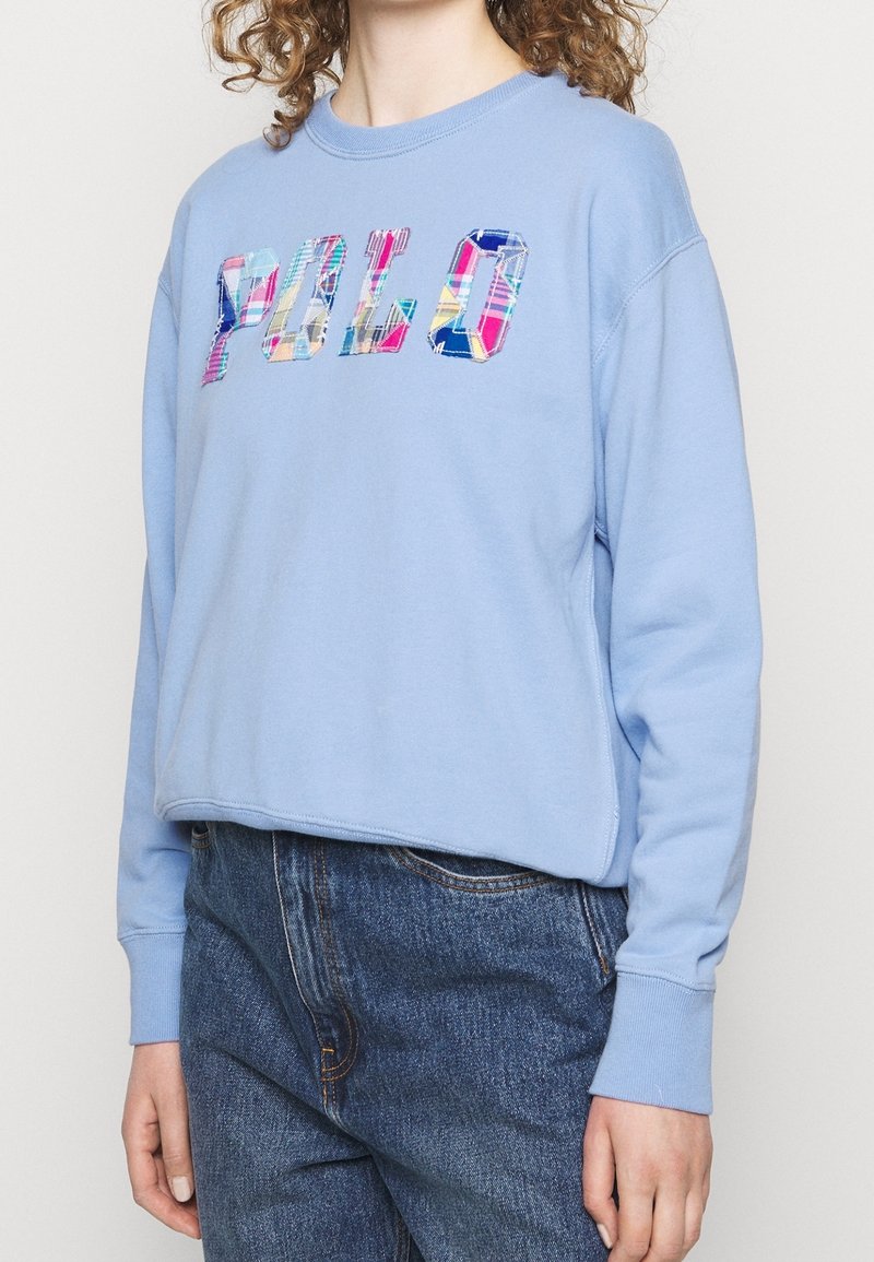 Personne portant un sweat-shirt bleu clair avec des lettres à carreaux colorés formant le mot « POLO » et un jean bleu taille haute.