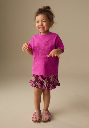 Smiling toddler standing, wearing a bright pink shirt with palm tree design, ruffled floral shorts, and pink sandals on a neutral background.