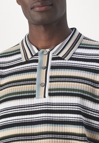 Homme portant un polo côtelé à rayures multicolores avec des lignes horizontales noires, blanches, beiges, vertes et bleu clair et trois boutons.