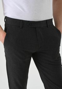 Black trousers made of textured fabric, featuring a flat front, side pockets, and a single button closure at the waist.