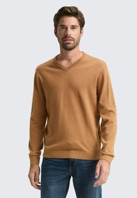 V-Ausschnitt, langärmeliger Pullover aus hellbraunem Stoff, mit strukturiertem Strick und gerippten Bündchen und Saum, kombiniert mit blauen Jeans.