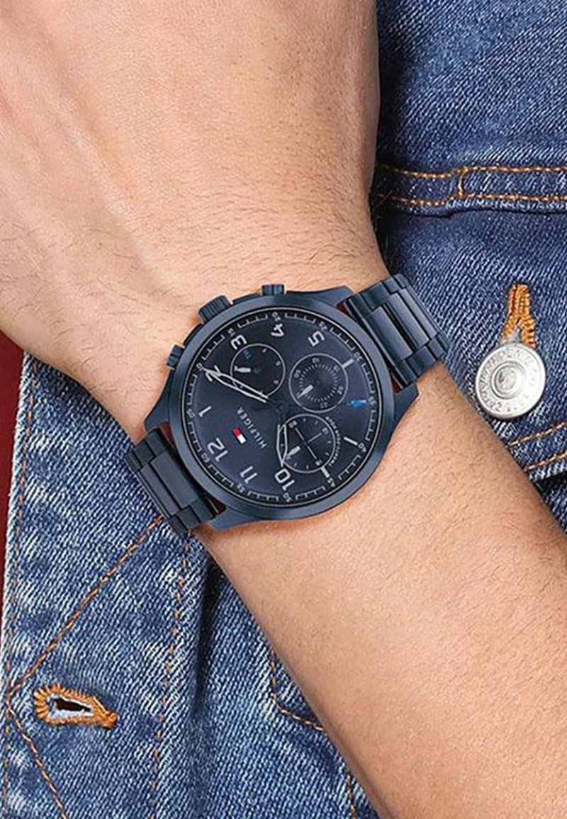 Montre chronographe noire avec cadran rond, bracelet en métal et quincaillerie argentée. Dispose de trois sous-cadrans et de marquages numériques proéminents.