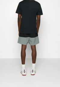 Muž stojící čelem k odvrácené straně, oblečený v černém tričku, dvoubarevných černobílých šortkách, bílých ponožkách a bílých teniskách s červenými podrážkami.