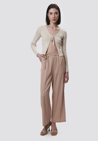 Helles beige, kurz geschnittenes Strickjäckchen mit Bindeband, kombiniert mit weit geschnittenen, beigen Hosen. Das Model trägt sandfarbene, strukturierte Absatzsandalen.