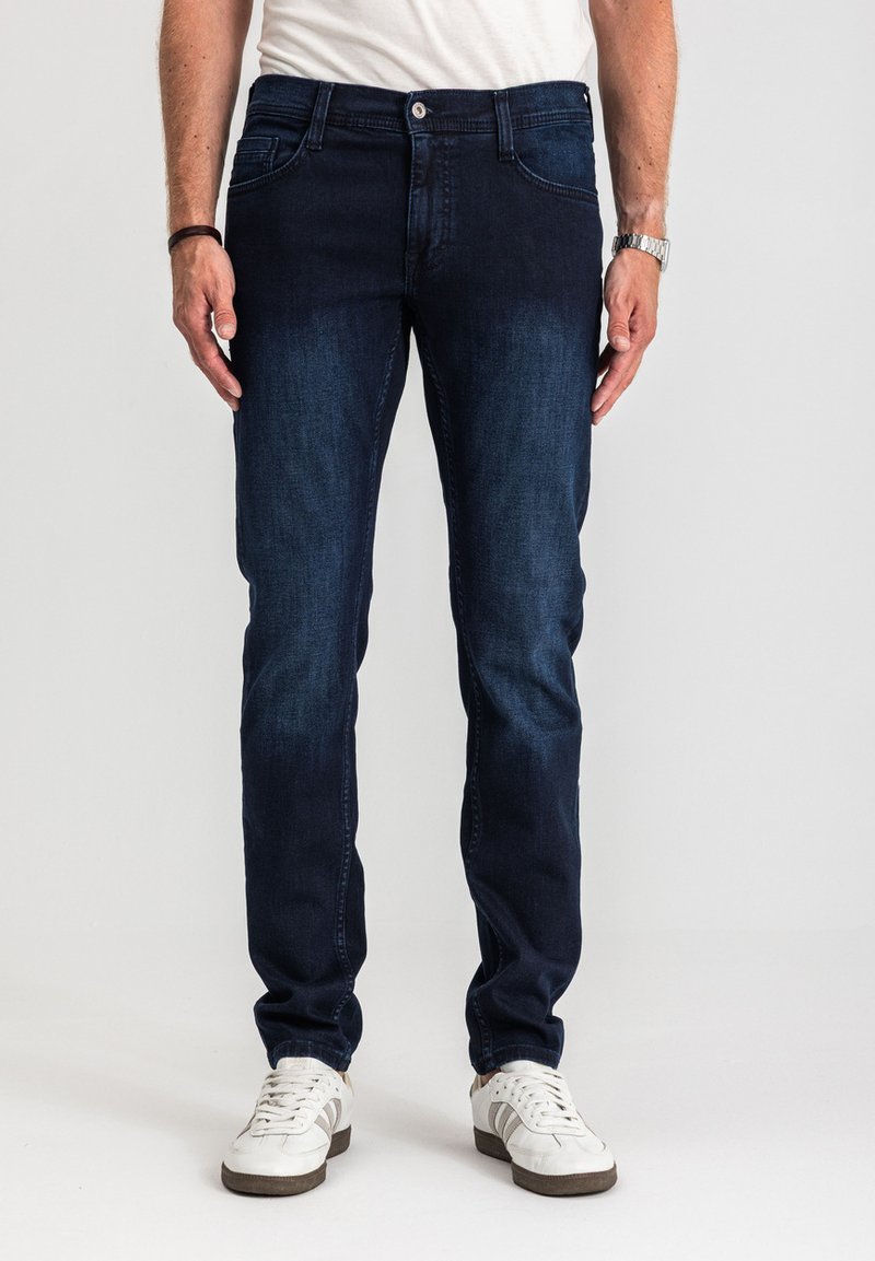 Mężczyzna ubrany w ciemnoniebieskie dopasowane jeansy, białe trampki, białą koszulkę, z zegarkiem na nadgarstku i bransoletką, stojący na neutralnym tle.