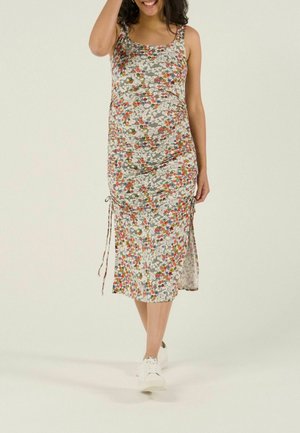 Lächelnde schwangere Frau, die einen ärmellosen, geblümten Midikleid mit Seitenschleifen und weißen Turnschuhen trägt, vor hellem Hintergrund läuft.