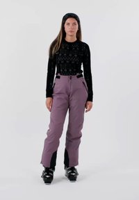 Pantalon de ski violet avec des accents noirs, bretelles ajustables, poches latérales et coupe profilée, associé à des bottes de ski noires.