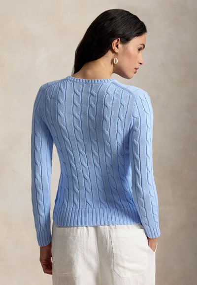 Vrouw die een lichtblauwe kabelgebreide trui en witte broek draagt, van achteren gezien met haar hoofd naar de zijkant gedraaid.