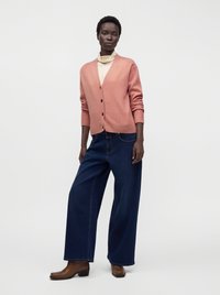 Femme noire debout portant un cardigan rose boutonné, un col roulé crème, un jean large bleu foncé et des bottines marron.