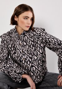 Quiltad blus i svart och vitt mönster med hög krage, guldknappar och puffärmar som har en geometrisk design och texturerad tyg.