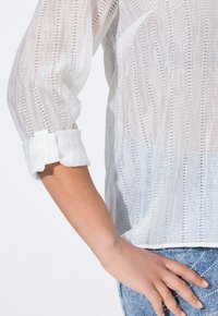 Blusa blanca semitransparente con rayas verticales, con mangas enrolladas y un tejido ligero y texturizado.