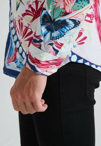 Chemise florale aux couleurs vives, avec un papillon bleu et des fleurs rouges. Tissu en coton à texture douce et détails de poignets boutonnés.