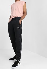 Schwarze Jogginghose mit weißen Seitenstreifen, elastischem Bund und Logo-Patch. Kombiniert mit einem hellrosa ärmellosen Oberteil und schwarzen Turnschuhen.