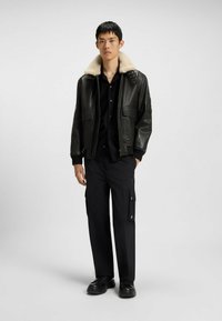 Czarna skórzana kurtka bomber z kremowym kołnierzem z owczej wełny, dwiema przednimi kieszeniami, długimi ściągaczami, w połączeniu z czarnymi spodniami cargo i czarnymi butami.