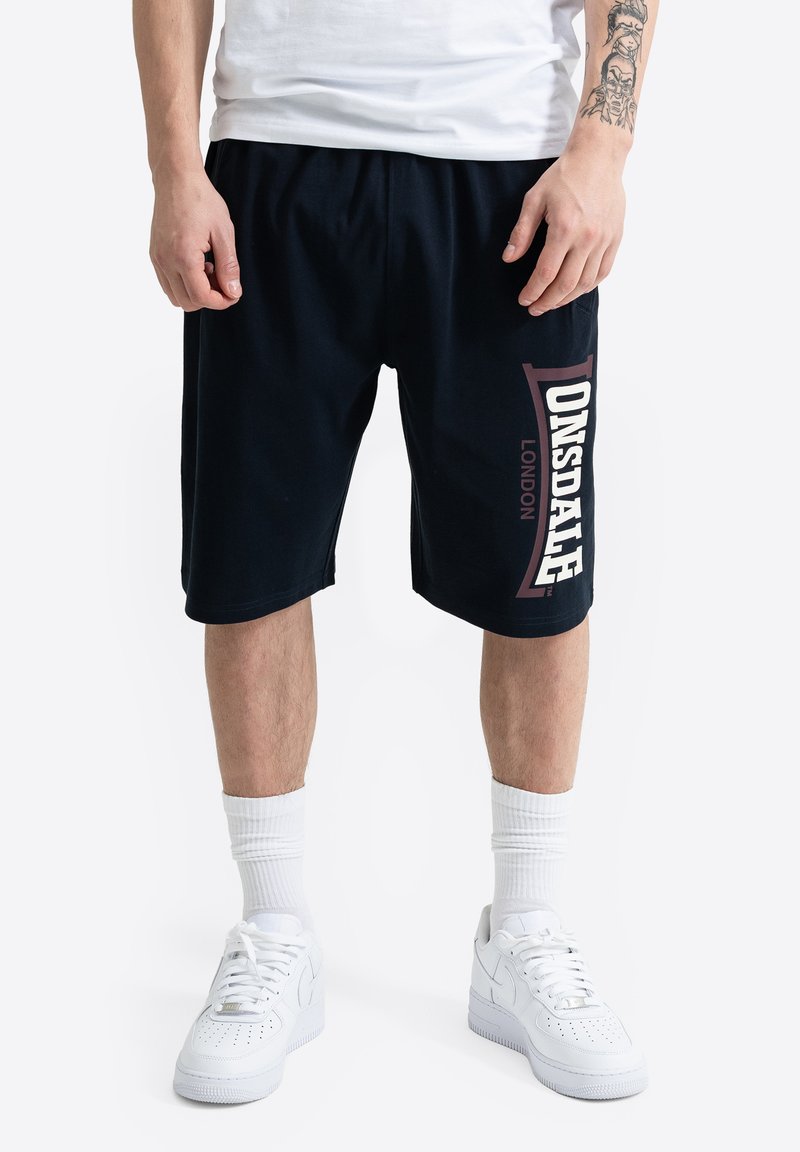 Άνδρας φορώντας μαύρα σορτς Lonsdale London, λευκές κάλτσες και λευκά αθλητικά παπούτσια, στέκεται με τα χέρια χαλαρά στα πλάγια μπροστά σε απλό φόντο.
