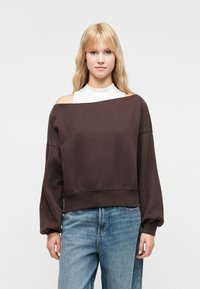 Sudadera marrón sin hombros con mangas caídas y dobladillo acanalado, combinada con una blusa blanca y jeans de un azul claro. Textura de tela suave.