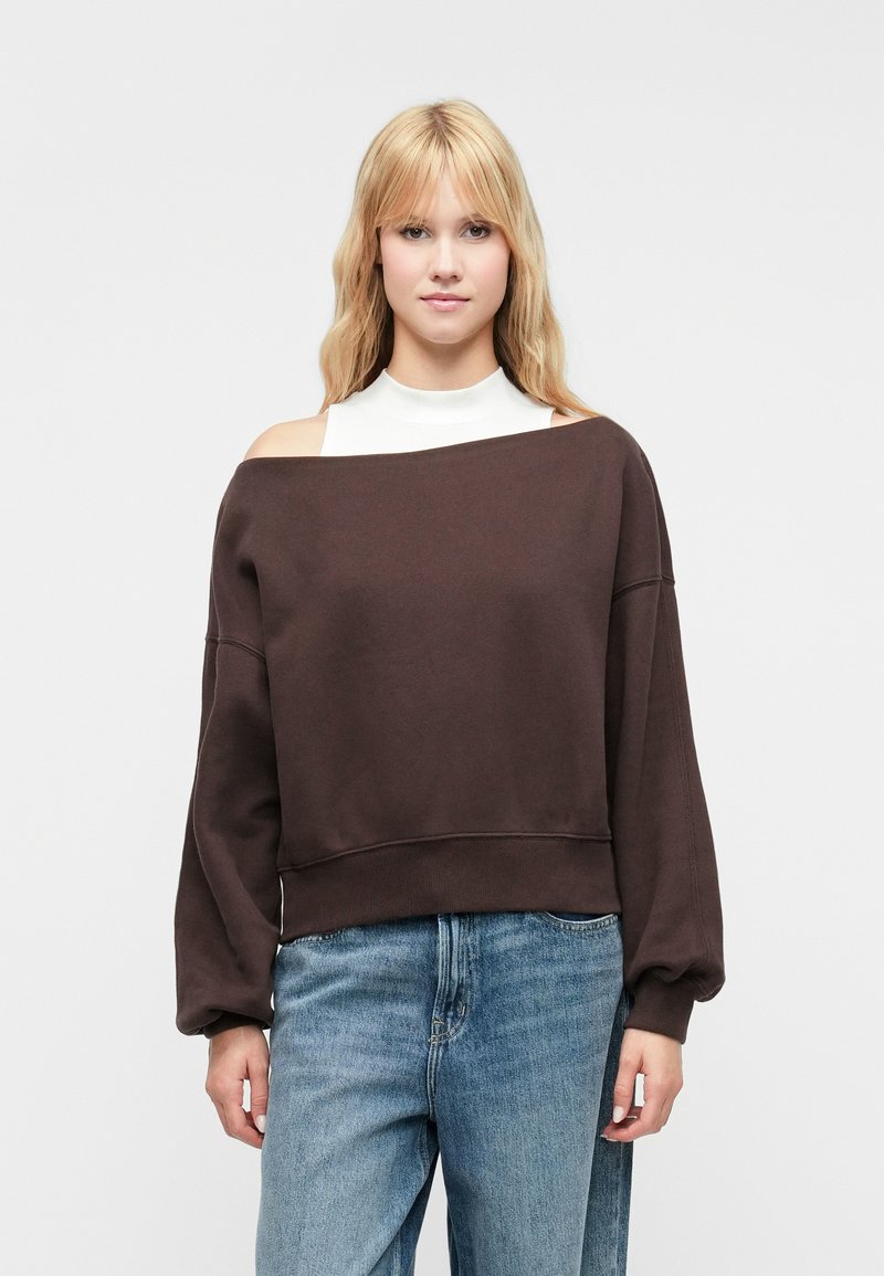 Sudadera marrón sin hombros con mangas caídas y dobladillo acanalado, combinada con una blusa blanca y jeans de un azul claro. Textura de tela suave.
